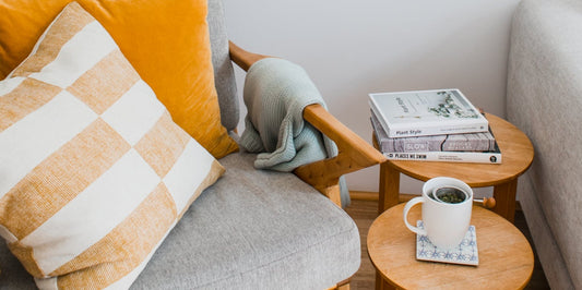 Living room side chair with two pillows and a blanket draped next to two side tables stacked with books and a coffee mug.